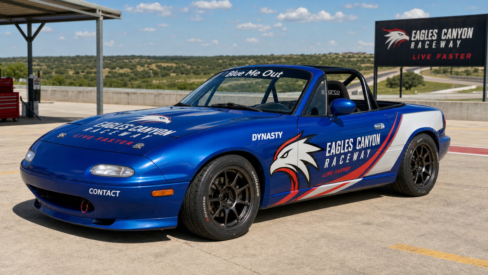 1990 Mazda Miata NA — Ethan's Build — Mariner Blue with full Live Faster Eagles Canyon Raceway livery wrap, eagle head graphic, Blue Me Out windshield banner, DYNASTY sponsor decal, Sparco racing seat, roll bar, black race wheels, photographed at Eagles Canyon paddock.