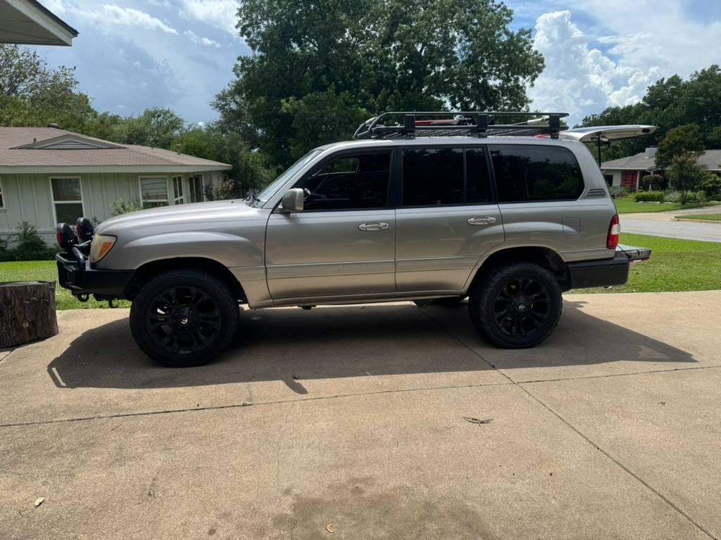 The Founder Truck — 2006 Toyota Land Cruiser 100-series in titanium silver, driver-side profile. ARB winch bumper with twin auxiliary lights, ARB rear bumper with jerry-can mount, full roof rack with Hi-Lift jack and ARB awning, lifted on Old Man Emu suspension, black aftermarket wheels with KO2 tires. Real truck, real driveway.